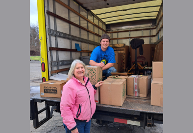 two people smiling for a photo at the back of an open truck surrounded by cardboard books of boxes