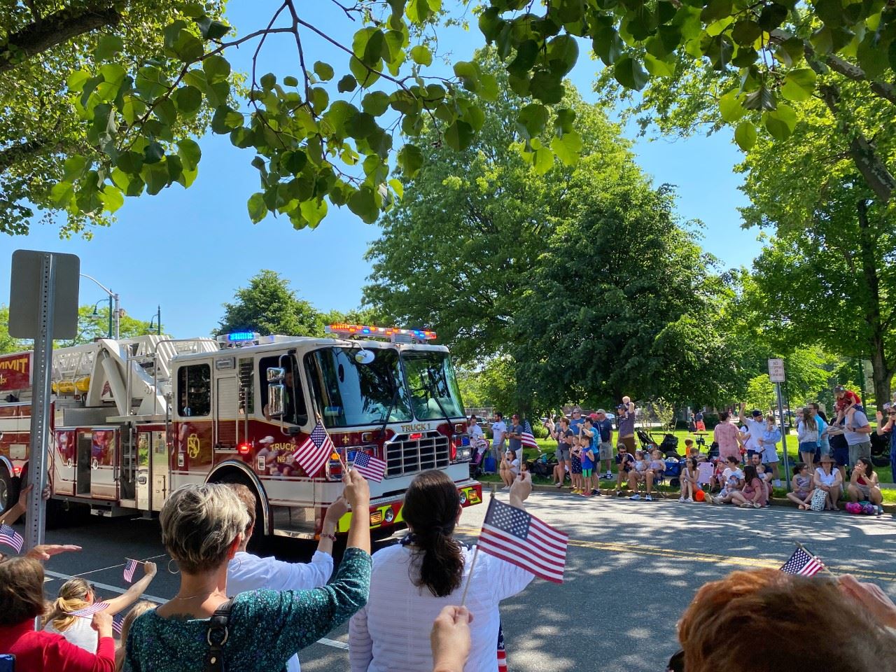image of firetruck driving down a street lined with people waving flags for memorial day